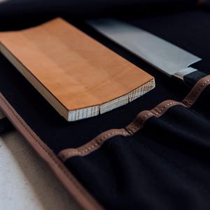 Puede incluir: Un rollo de cuchillos de cuero marrón y lona negra con una tabla de cortar de madera y un cuchillo plateado en su interior.