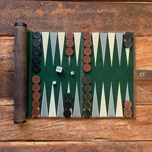 May include: A green felt backgammon board with black and brown playing pieces. The board is rolled up and has a brown leather carrying case. There are two dice on the board, one showing a 4 and the other showing a 3.