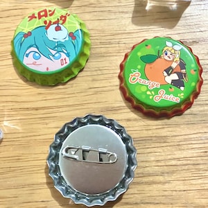 May include: Three bottle caps with different designs. The first cap features a green background with a cartoon girl and the text "メロンソーダ" in Japanese. The second cap features a green background with a cartoon girl holding an orange and the text "Orange Juice". The third cap is silver and has a safety pin attached to the back.