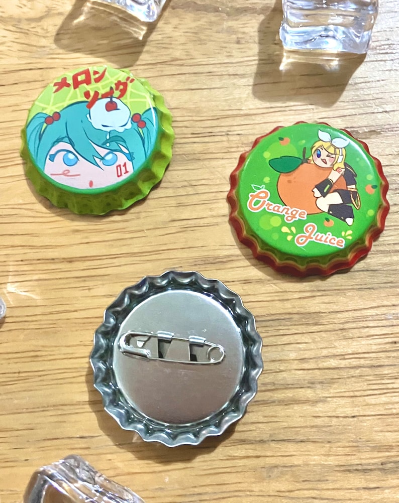 Puede incluir: Tres tapones de botella con diferentes dise&ntilde;os. El primer tap&oacute;n tiene un fondo verde con una ni&ntilde;a de dibujos animados y el texto "メロンソーダ" en japon&eacute;s. El segundo tap&oacute;n tiene un fondo verde con una ni&ntilde;a de dibujos animados sosteniendo una naranja y el texto "Orange Juice". El tercer tap&oacute;n es plateado y tiene un pasador de seguridad en la parte posterior.