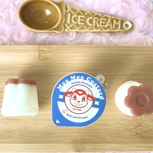 May include: A wooden tray with three miniature desserts: a white and pink popsicle, a white and brown pudding cup with a flower on top, and a blue and red circular sticker with the text "Moo Moo Custard, Vanilla Flavored! Est. 1998, Rich and Creamy!"