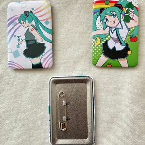 May include: Three rectangular pinback buttons featuring anime-style illustrations. Two buttons display a teal-haired character in different poses against colorful backgrounds. The third button is a metal pinback with a safety pin attached.