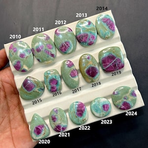 May include: A selection of polished gemstones in various shapes, showcasing a mix of green and purple colours. Some stones have a marbled appearance. The gemstones are displayed on a white surface, with each stone labelled with a number.