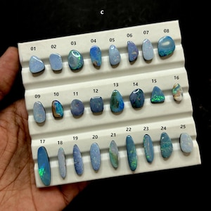 Könnte beinhalten: Eine Sammlung von 25 polierten Opal-Edelsteinen in verschiedenen Formen und Größen, präsentiert auf einem weißen Tablett. Die Opale zeigen eine Reihe von Farben, darunter Blau, Grün und schillernde Blitze. Jeder Stein ist nummeriert.