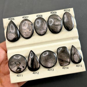 Pode incluir: Uma coleção de pedras preciosas de obsidiana polida, cinza escuro, em várias formas, incluindo oval, gota e redonda. Cada pedra tem um padrão único e é numerada. As pedras preciosas são exibidas em uma superfície branca.