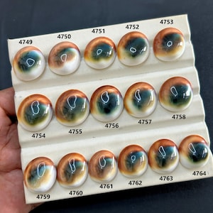 May include: A selection of polished, round gemstones with an eye-like design. The stones display a mix of brown, green, and white hues, with a shiny surface. They are arranged on a white display card, each stone labelled with a number. Measurements are not available.