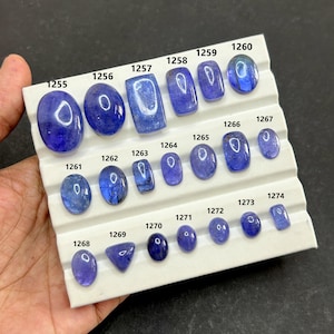 May include: A collection of polished, blue gemstones of various shapes and sizes, displayed on a white tray. Each stone is numbered, ranging from oval to rectangular cuts. The gemstones are held by a hand.