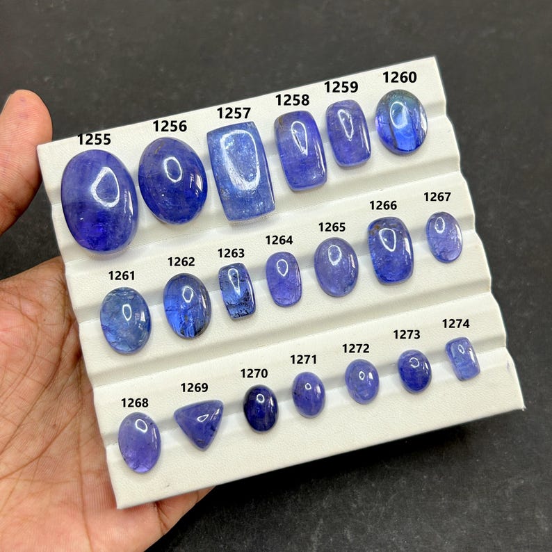 May include: A collection of polished, blue gemstones of various shapes and sizes, displayed on a white tray. Each stone is numbered, ranging from oval to rectangular cuts. The gemstones are held by a hand.
