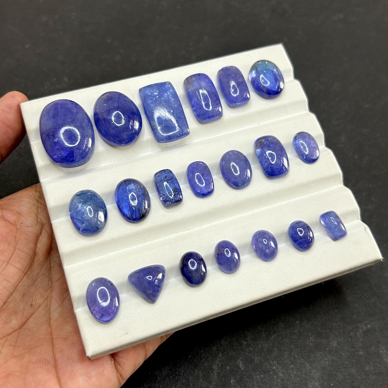 May include: An assortment of polished, blue-violet gemstones in various shapes and sizes, including ovals, rectangles, and a triangle, are displayed on a white, tiered tray. The gemstones have a glossy finish and are arranged in neat rows.