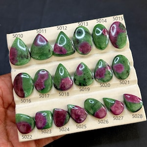 Puede incluir: Una colección de piedras preciosas de rubí en zoisita pulidas, cada una con una forma única y una mezcla de colores verde y magenta. Las piedras están dispuestas sobre una tarjeta blanca, con números individuales visibles.