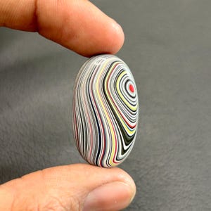 May include: A colorful agate cabochon with a swirling pattern of white, black, red, yellow, and blue lines. The cabochon is oval shaped and has a smooth, polished surface.