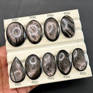 Pode incluir: Uma coleção de pedras preciosas polidas, em forma oval, com um brilho metálico, exibidas em uma superfície branca. As pedras variam em tamanho e têm uma aparência escura e reflexiva com padrões únicos. Cada pedra é numerada.