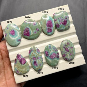 May include: A collection of eight polished gemstones, each with a unique shape. The stones feature a mottled pattern of teal and magenta hues. Each stone is numbered with a white label. The gemstones are displayed on a white tray.