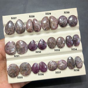 Peut inclure: Collection de pierres précieuses roses et brunes, taillées en formes variées, dont des ovales et des gouttes. Les pierres sont présentées sur un présentoir blanc, chacune numérotée. Les facettes des pierres révèlent leurs motifs naturels.