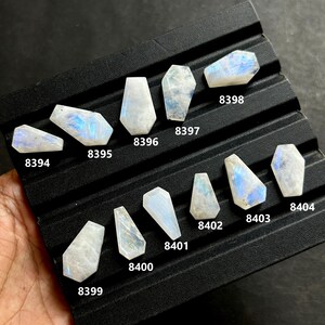 May include: A collection of polished, iridescent moonstone gemstones in various shapes, including coffin and trapezoid cuts. The stones display a range of colors, including white, blue, and flashes of rainbow hues. Each stone is labeled with a number.