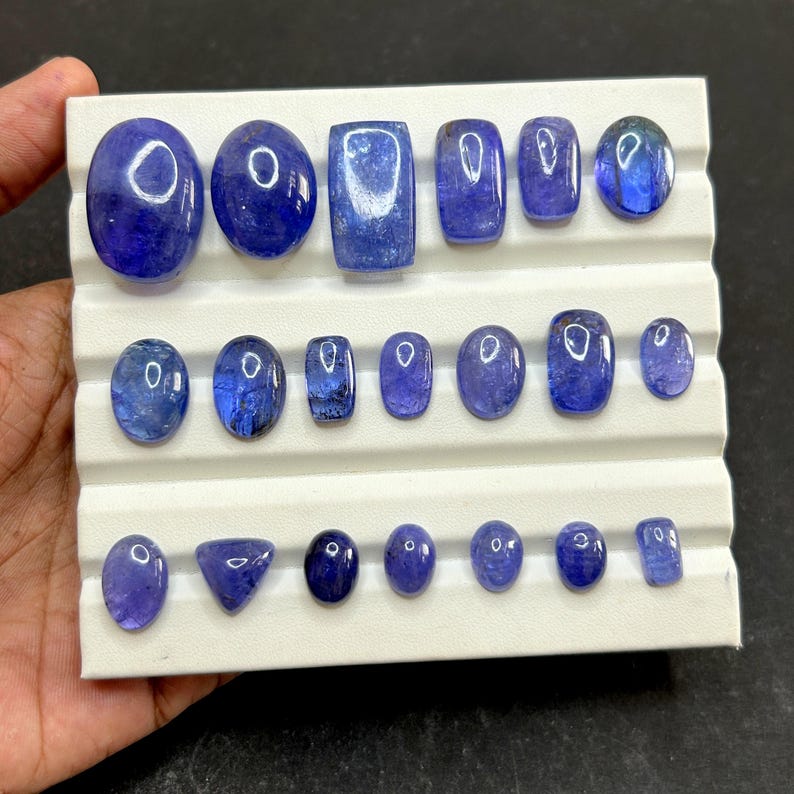 May include: A collection of polished, blue-violet gemstones in various shapes, including ovals, rectangles, and a triangle, arranged on a white display tray. The gemstones have a translucent quality, reflecting light.