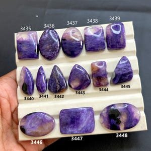 Puede incluir: Una colección de piedras preciosas de charoíta pulidas en varias formas, incluyendo rectángulos, óvalos y lágrimas. Las piedras muestran una gama de tonos púrpuras con patrones arremolinados y algunas con inclusiones marrones y blancas. Cada piedra está numerada.