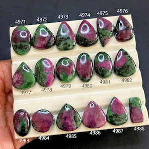 Pode incluir: Uma coleção de pedras preciosas de rubi zoisite em várias formas, incluindo ovais, em forma de gota e de pêra. As pedras exibem uma mistura de tons vermelho escuro e verde, com inclusões visíveis. Cada pedra está numerada.