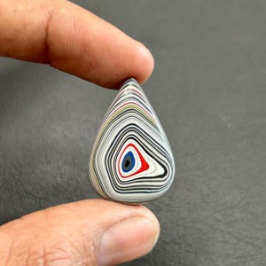 Puede incluir: Un cabujón de ágata en forma de lágrima con un patrón de remolino de negro, blanco, rojo, azul y amarillo. El ágata tiene un patrón central en forma de ojo.