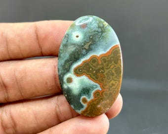 Op de afbeelding: Een ovaalvormige edelsteen met een gepolijst oppervlak. De steen heeft een mix van kleuren, waaronder groen, bruin en oranje, met een uniek patroon. De edelsteen wordt in een hand gehouden, wat de grootte en vorm benadrukt.