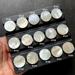 Può includere: Una collezione di perline rotonde di madreperla iridescente, ognuna con un motivo unico e numerata da 8148 a 8162. Le perline sono esposte su un vassoio nero, mostrando la loro lucentezza naturale e le variazioni di colore.