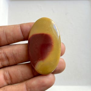 Puede incluir: Piedra pulida de forma ovalada con una superficie lisa y brillante. La piedra presenta un llamativo patrón de color, con una gran sección de color rojo intenso sobre un fondo amarillo dorado. La piedra se sostiene en una mano.