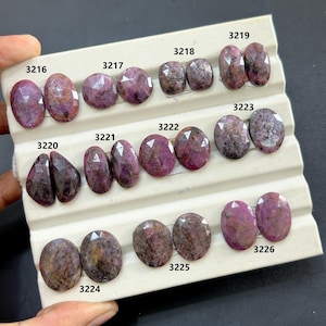 May include: A collection of faceted, oval-shaped gemstones in varying shades of red and purple. Each pair of stones is labeled with a number. The gemstones are displayed on a white, textured surface, showcasing their polished surfaces and internal patterns.