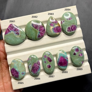 May include: A collection of nine polished gemstones, each with a unique shape and pattern. The stones feature a mix of colors, including a dominant green hue with vibrant red and pink inclusions. The gemstones are displayed on a white tray.