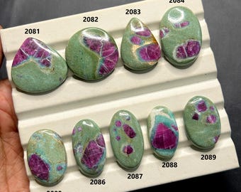 Può includere: Una collezione di nove pietre preziose lucide, ognuna con una forma e un motivo unici. Le pietre presentano un mix di colori, tra cui una tonalità verde dominante con inclusioni rosse e rosa vibranti. Le pietre sono esposte su un vassoio bianco.