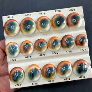 Pode incluir: Uma coleção de pedras preciosas cabochão polidas, cada uma com um padrão único semelhante a um olho. As pedras apresentam uma mistura de cores, incluindo marrom, verde e branco, e estão dispostas em um cartão de exibição branco. Cada pedra é numerada.