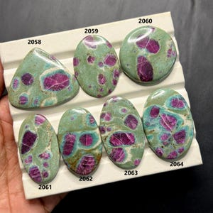 May include: A collection of polished gemstones with a green matrix and vibrant magenta inclusions. The stones vary in shape, including oval, round, and triangular. Each stone has a unique pattern of color and texture, showcasing natural variations.