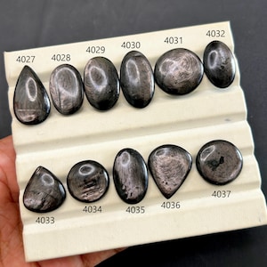 Pode incluir: Uma coleção de pedras preciosas pretas polidas com veios prateados iridescentes, exibidas numa bandeja branca. As pedras variam em forma, incluindo oval, gota e redonda, e estão numeradas. As pedras preciosas são provavelmente para joalharia ou colecionismo.