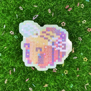 May include: A pixelated, iridescent sticker in the shape of a drink with a whipped cream topping. The sticker has a light-colored border and is surrounded by green grass and scattered star-shaped confetti.