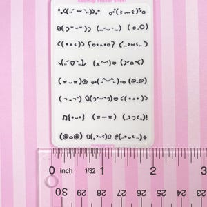 May include: A white sticker sheet with various kaomoji emoticons. The sheet is approximately 2 inches long, as measured by the ruler. The text "kaomoji sticker sheet" is printed at the top. The background is pink and white striped.