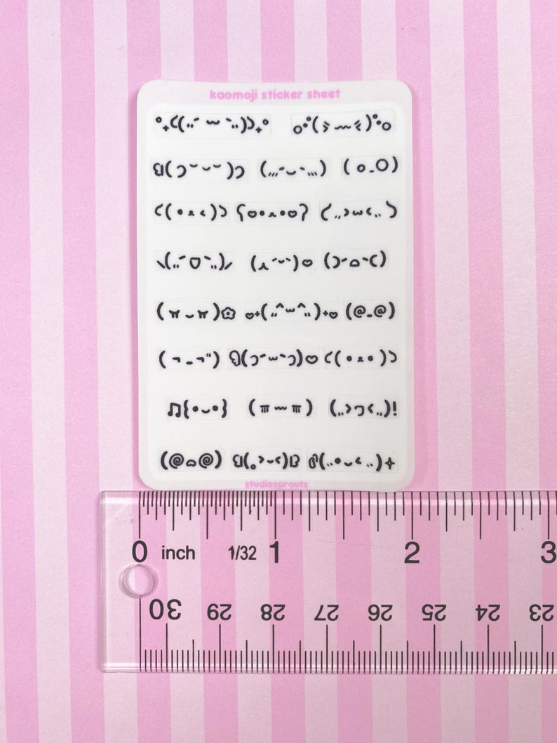 May include: A white sticker sheet with various kaomoji emoticons. The sheet is approximately 2 inches long, as measured by the ruler. The text "kaomoji sticker sheet" is printed at the top. The background is pink and white striped.