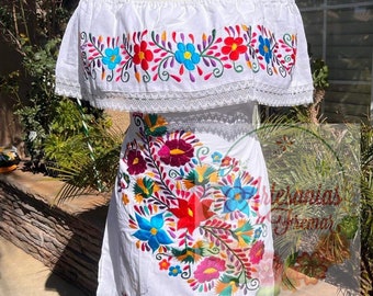 May include: White off-the-shoulder dress with vibrant floral embroidery in red, blue, yellow, and purple. The dress features a ruffled neckline and hem, showcasing a traditional Mexican design. The embroidery is detailed and colorful, creating a festive look.