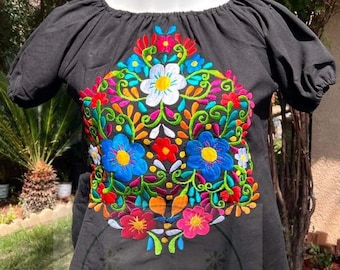 May include: Black cotton blouse with short sleeves and a floral embroidery design in shades of pink, blue, green, yellow, and orange. The embroidery features a large floral motif with a scalloped lace trim at the bottom.