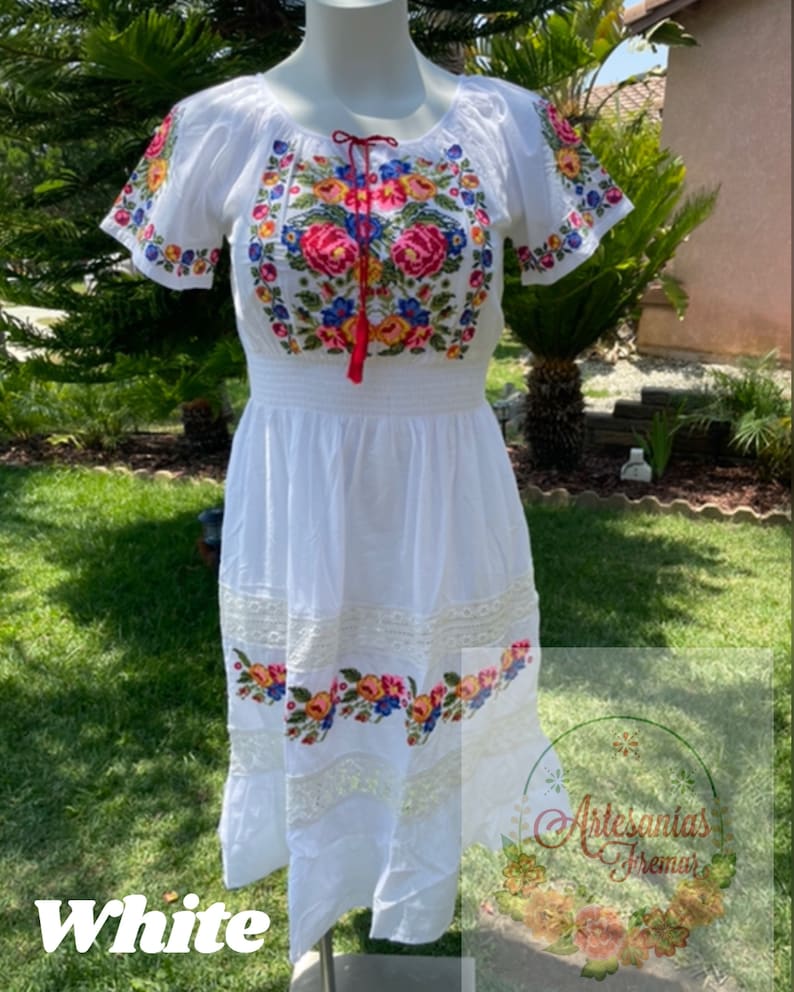 May include: White embroidered cotton dress with floral designs and a lace trim. The dress has a gathered waist and short sleeves. The text "Artesanias Iremar" is visible on the bottom right corner of the image.
