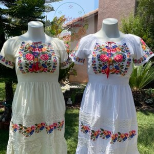 May include: Two white embroidered dresses with floral patterns. The dress on the left is cream colored and the dress on the right is white. Both dresses have short sleeves and a gathered waist.
