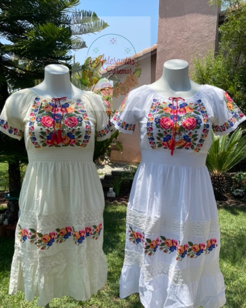 May include: Two white embroidered dresses with floral patterns. The dress on the left is cream colored and the dress on the right is white. Both dresses have short sleeves and a gathered waist.