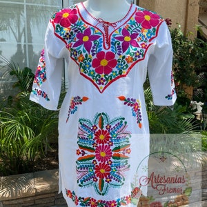 May include: White cotton tunic with colorful floral embroidery. The embroidery features pink, red, orange, yellow, green, blue, and purple flowers. The text "Artesanias Fremar" is embroidered on the bottom right corner of the tunic.