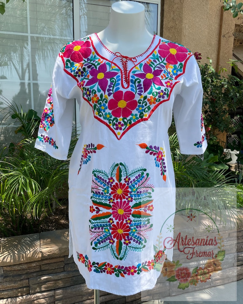 May include: White cotton tunic with colorful floral embroidery. The embroidery features pink, red, orange, yellow, green, blue, and purple flowers. The text "Artesanias Fremar" is embroidered on the bottom right corner of the tunic.