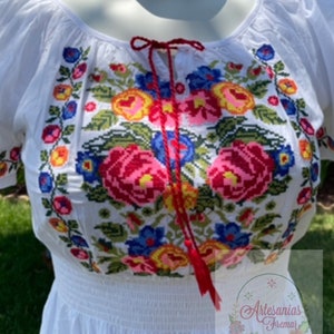 May include: White embroidered blouse with a red ribbon tie and a floral pattern in shades of red, pink, yellow, blue, and green. The blouse has a gathered elastic waistband.
