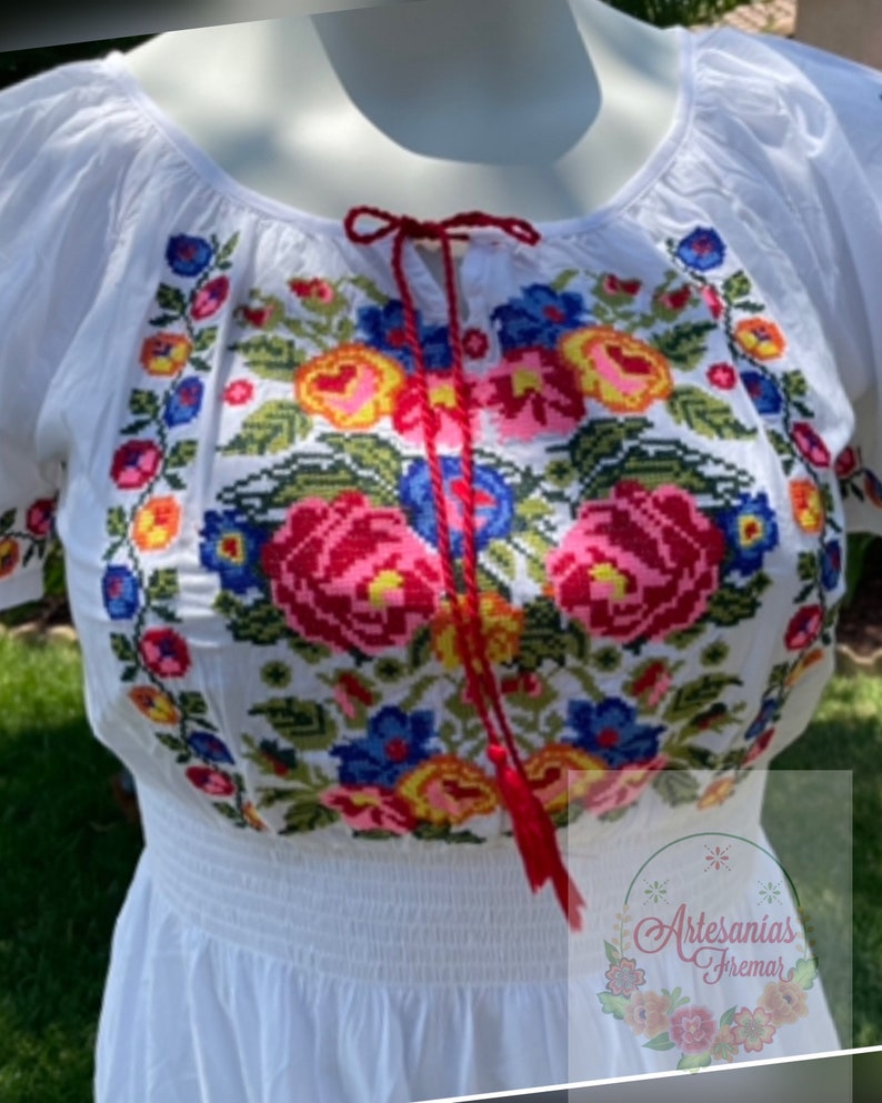 May include: White embroidered blouse with a red ribbon tie and a floral pattern in shades of red, pink, yellow, blue, and green. The blouse has a gathered elastic waistband.