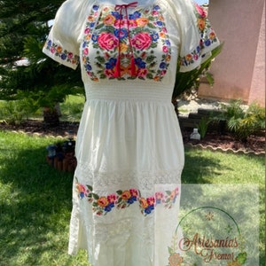 May include: A white embroidered dress with a floral design. The dress has short sleeves and a fitted bodice. The skirt is flowy and has a lace trim. The dress is made of cotton and is perfect for summer.
