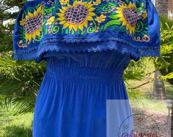 May include: A blue off-the-shoulder top with a sunflower embroidery design. The top has a ruffled neckline and a fitted bodice. The text "Artesanías Iremar" is visible in the lower right corner of the image.