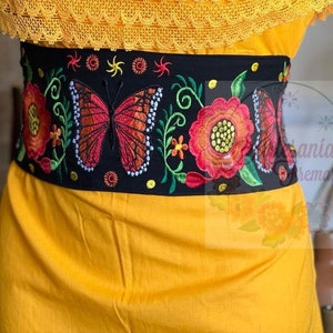 May include: A black embroidered belt with a large butterfly design in red, orange, and yellow. The belt is decorated with floral motifs and has a black background. The belt is worn over a yellow dress.
