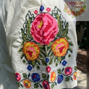 May include: White cotton blouse with embroidered floral details in shades of pink, yellow, green, and blue. The embroidery features a large rose with smaller flowers and leaves surrounding it.