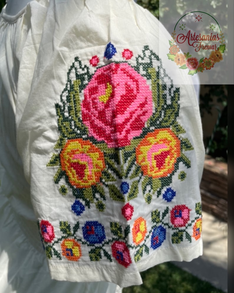 May include: White cotton blouse with embroidered floral details in shades of pink, yellow, green, and blue. The embroidery features a large rose with smaller flowers and leaves surrounding it.