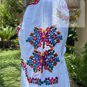 May include: White embroidered blouse with colorful floral designs. The blouse has a tie at the waist and three-quarter sleeves.